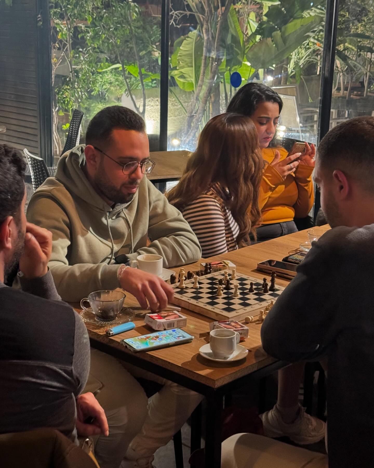 A chess game on a wooden table, coffees beside it, in the evening
